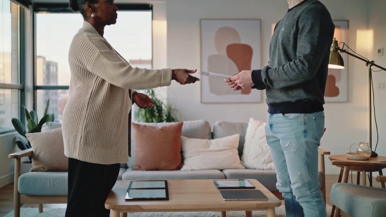 Femme en cardigan tendant un document, homme prenant le papier pour examen à la maison, avec des tablettes.