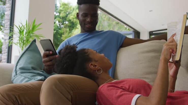 Video of happy african american couple lying on sofa with smartphone and book