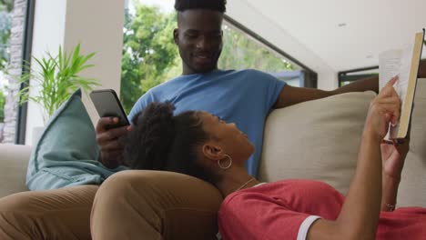 Video-of-happy-african-american-couple-lying-on-sofa-with-smartphone-and-book