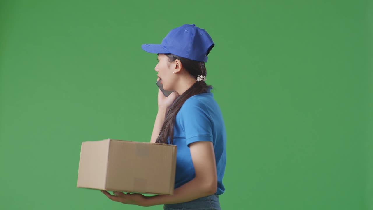 Side View Of Asian Female Courier In Blue Uniform Walking And Talking On Smartphone While Delivering A Carton On Green Screen Background In The Studio
