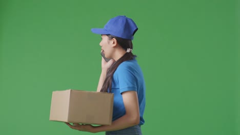vista lateral de una mensajera asiática en uniforme azul caminando y hablando en un teléfono inteligente mientras entrega una caja de cartón en el fondo de la pantalla verde en el estudio