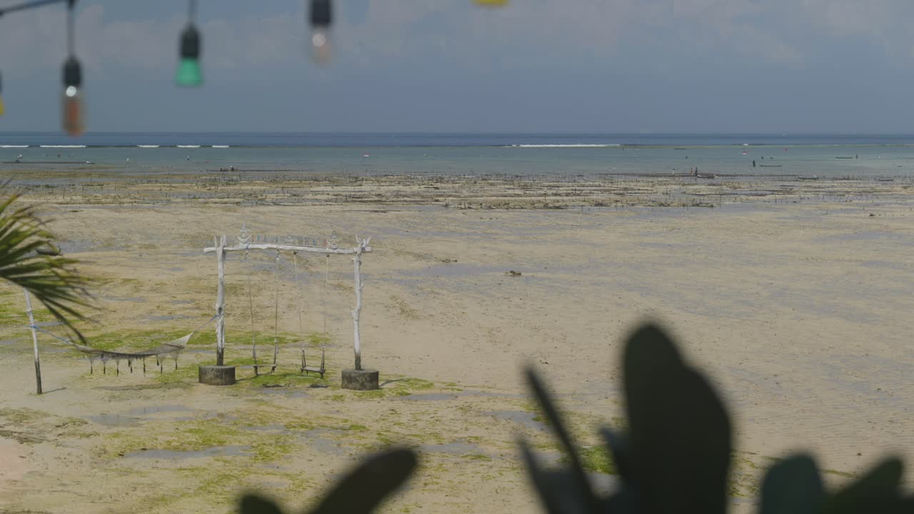 Looking out past party lights to low tide between Ceningan and Lembongan Islands off Bali, swings and hammocks sit above the water at high tide wide shot