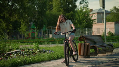 cyclist mounts bicycle near vibrant garden, adjusting hair under warm sunlight, surrounded by colorful flower beds, trees, and wooden bench, she prepares for an outdoor ride