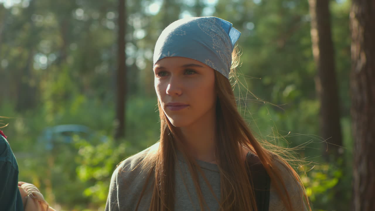 Lady wearing blue headscarf, lost in thought as her hair gently flutters in wind, she stands in sunlit forest with tall trees in background, partially accompanied by another person nearby