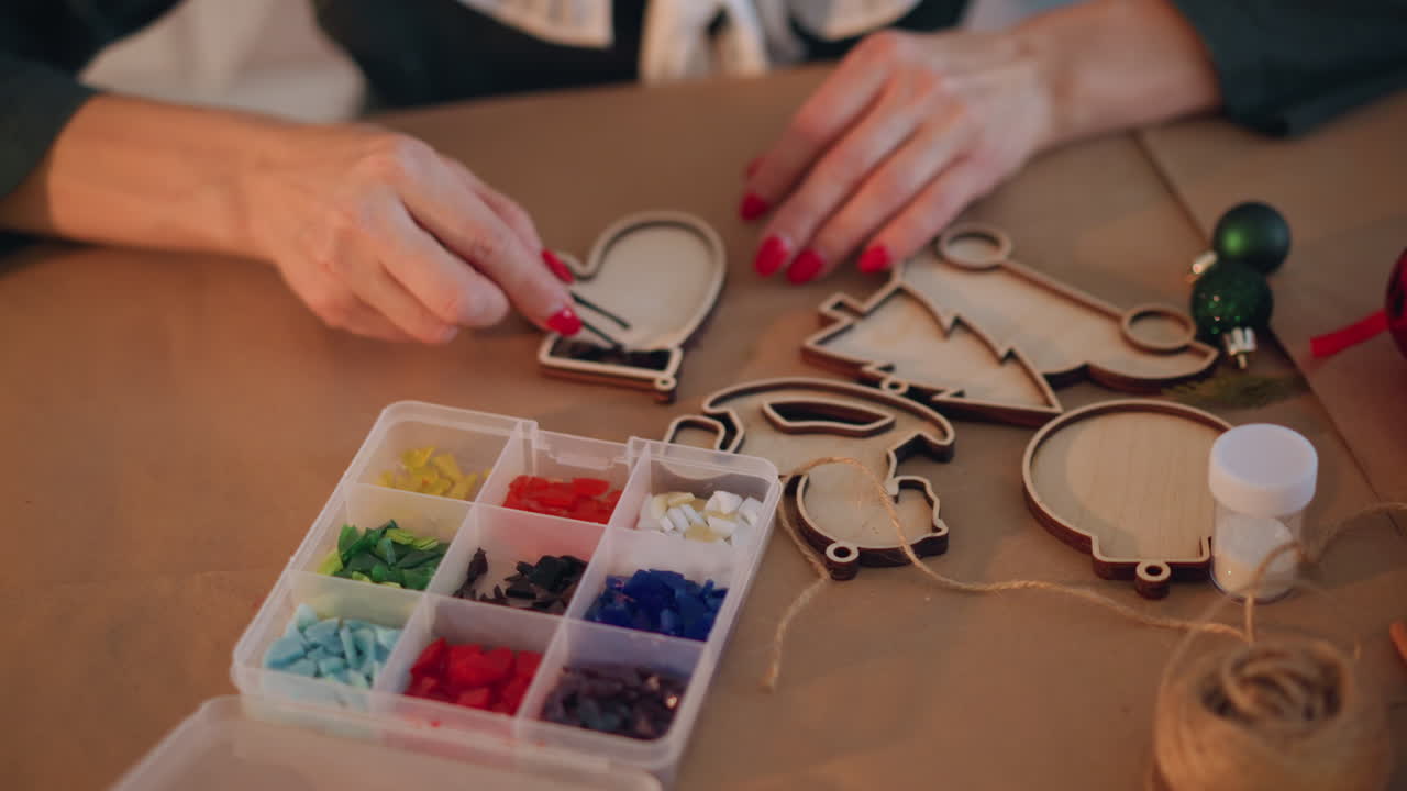 Closeup hands crafting wooden christmas ornaments on table with beads, ribbon, glue female with red manicure placing colorful beads into ornament frames warm festive lighting, cozy holiday DIY mood