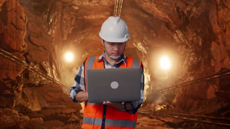 asian male engineer with safety helmet working on a laptop while standing in underground mine tunnel