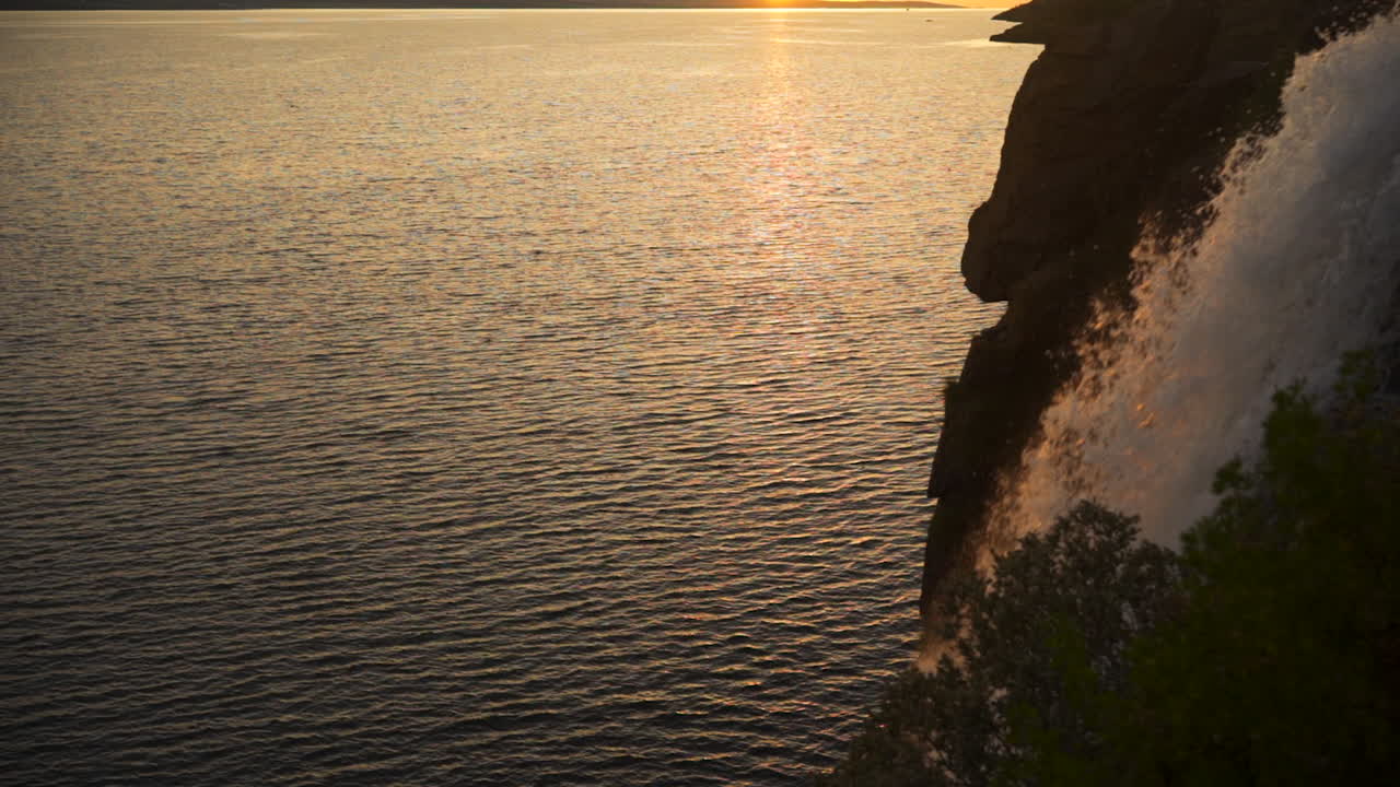 Golden hour sunset light illuminates ocean water in fjord and casts glow on waterfall cascade, slow motion