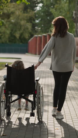 woman walks on stone walkway moving wheelchair with little daughter. mother takes care of girl with cerebral palsy spending time in park backside view