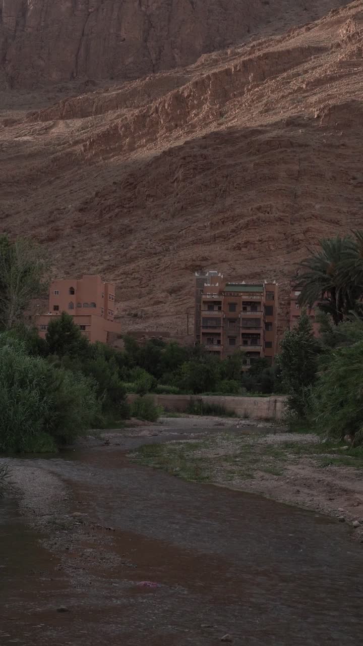 Un pendio montuoso desertico con un piccolo fiume con una portata d'acqua molto bassa. Si possono notare alcuni edifici con della vegetazione.