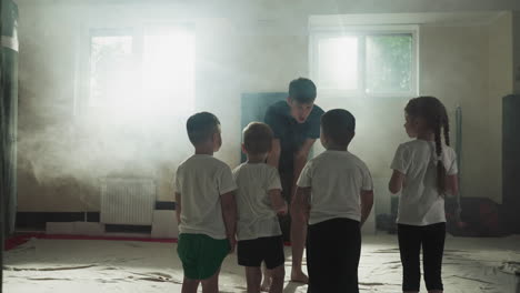 young trainer and children group in martial arts gym class. curious little kids look at sensei before exercising in misty gym slow motion. junior sportsmen