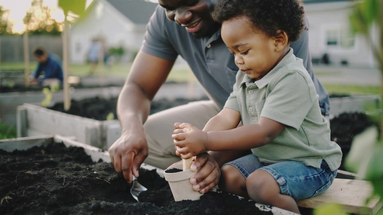 Reaching father guiding toddler filling small pot at raised bed, using trowel and dibber to plant