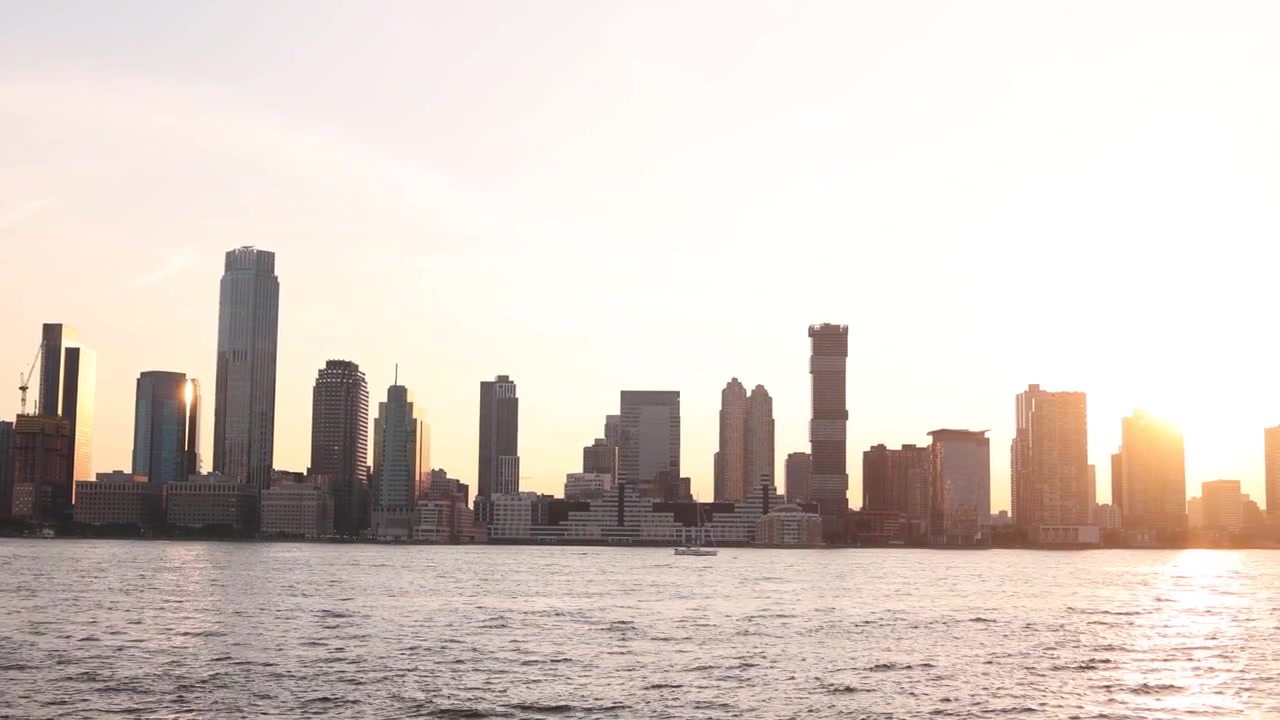 Panorâmica aérea ampla do horizonte de Nova York silhuetado durante um pôr do sol vibrante. Os arranha-céus da metrópole refletem a luz do entardecer que se esvai sobre o Rio Hudson.