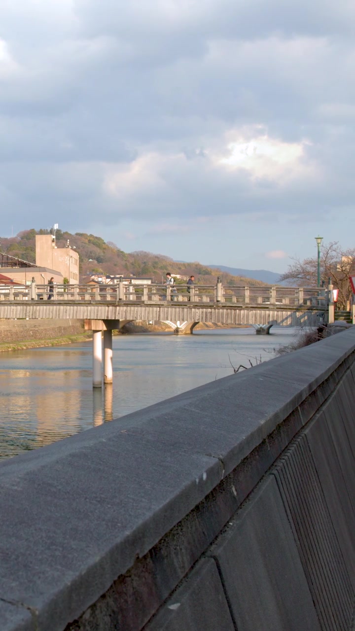 Ampia inquadratura fissa di un ponte tradizionale e architettura sulla riva del fiume sotto un cielo nuvoloso.