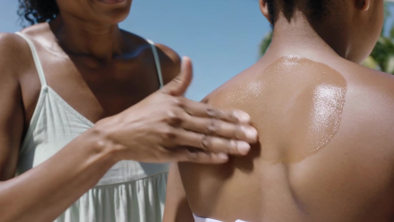 Spraying mom in dress rubbing sunscreen onto girl at beach sun shining, white bottle, strapless top