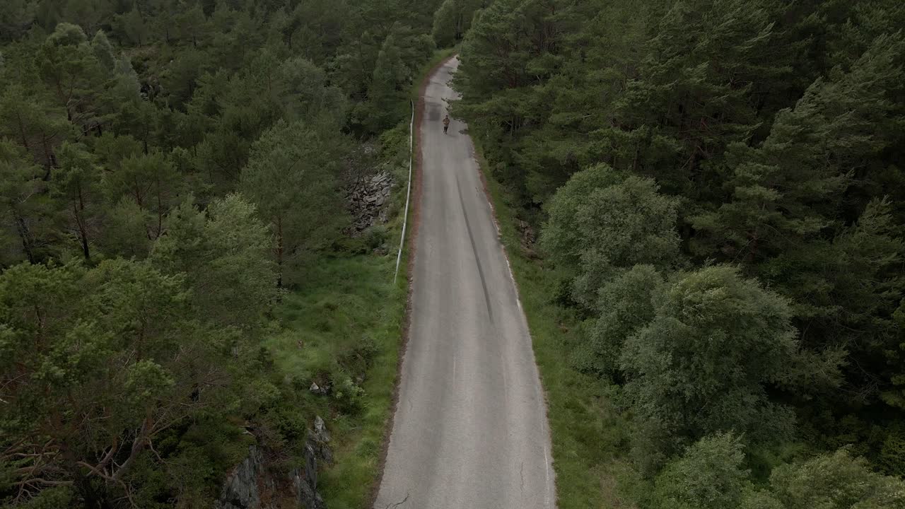 Aerial top down shot of person jogging on road between blowing forest trees on grey day in Norway