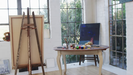 Close-up-of-canvas-on-easel-and-paints-and-brushes-on-table-in-studio,-slow-motion