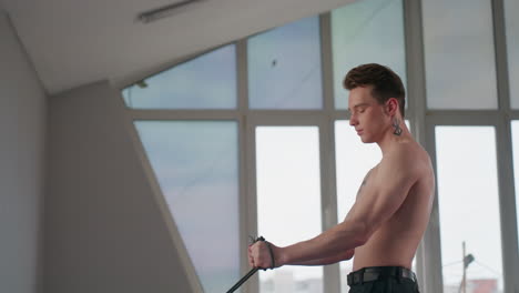 shirtless young man with neck tattoo stands, grips heavy resistance rope handles, braces core and pulls rope downward in focused muscle building workout against city window backdrop