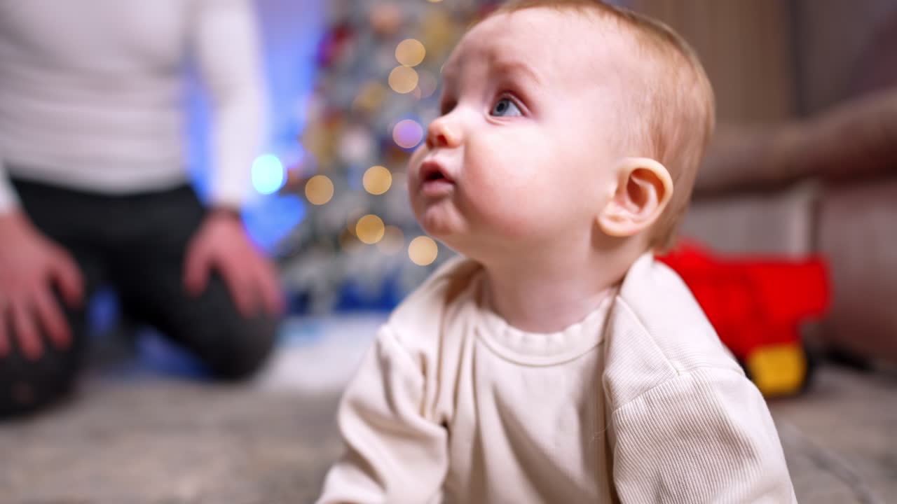 Adorable little toddler crawls by the floor approaching camera. A red cake is shown to baby, distracting him for a moment. Close up. Unrecognized man sits on the floor at backdrop.