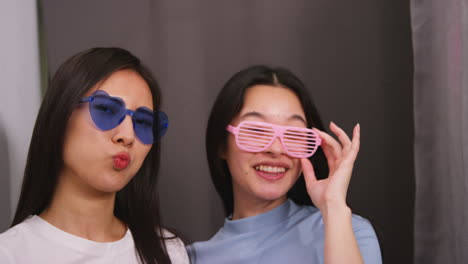 two female friends in photo booth wearing cool glasses having fun posing for portrait and pulling faces in real time