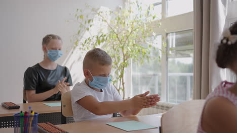 teacher children with face mask at school after lockdown disinfecting hands. school boy holding out his hands while sitting at desk in classroom as female teacher pouring some sanitize