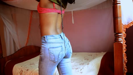 african woman taking off cropped belly shirt revealing pink bra wearing tight jeans in front of a bed in a hotel room - side view