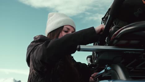 a millennial female, bundled in cute winter gear, unhooks bungy netting from the top of an suv roof rack basket