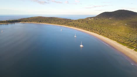 Playa-Frente-A-La-Bahía-De-Prueba-Con-Veleros-Navegando-En-El-Mar-Azul-Al-Atardecer---Exuberante-Montaña-Verde-Y-Bosque-En-El-Parque-Nacional-Arakoon-En-Nsw,-Australia