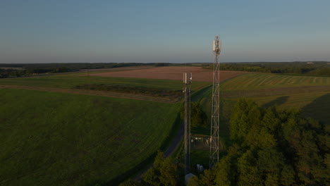 Two-radio-towers-in-the-middle-of-farmland-during-sunrise,-aerial-orbital-dolly-in-tilting