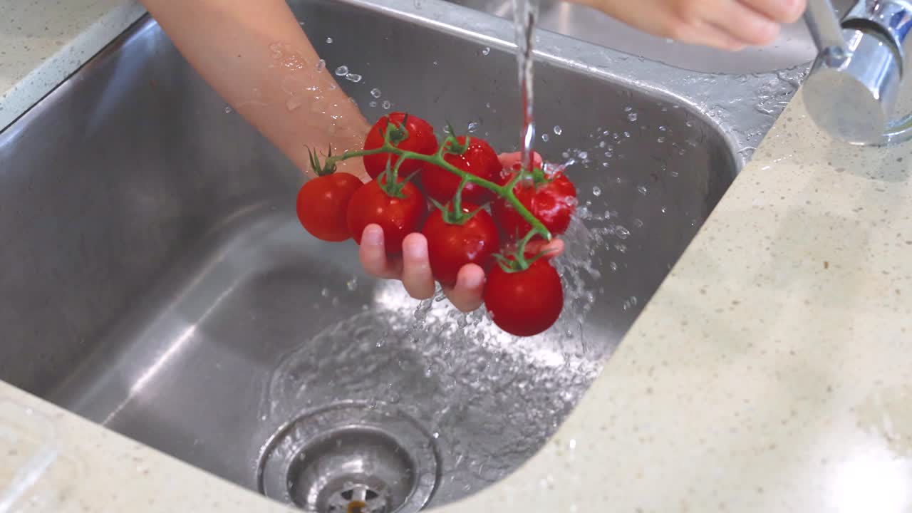 Hands washing vine tomatoes under a kitchen faucet. Bright lighting, close-up view, emphasizing cleanliness and freshness