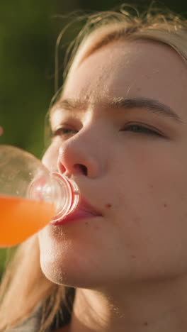 mujer al aire libre bebiendo jugo de naranja de una botella de plástico con contenido y expresión relajada, rodeada de naturaleza y luz solar suave, con vegetación borrosa en el fondo