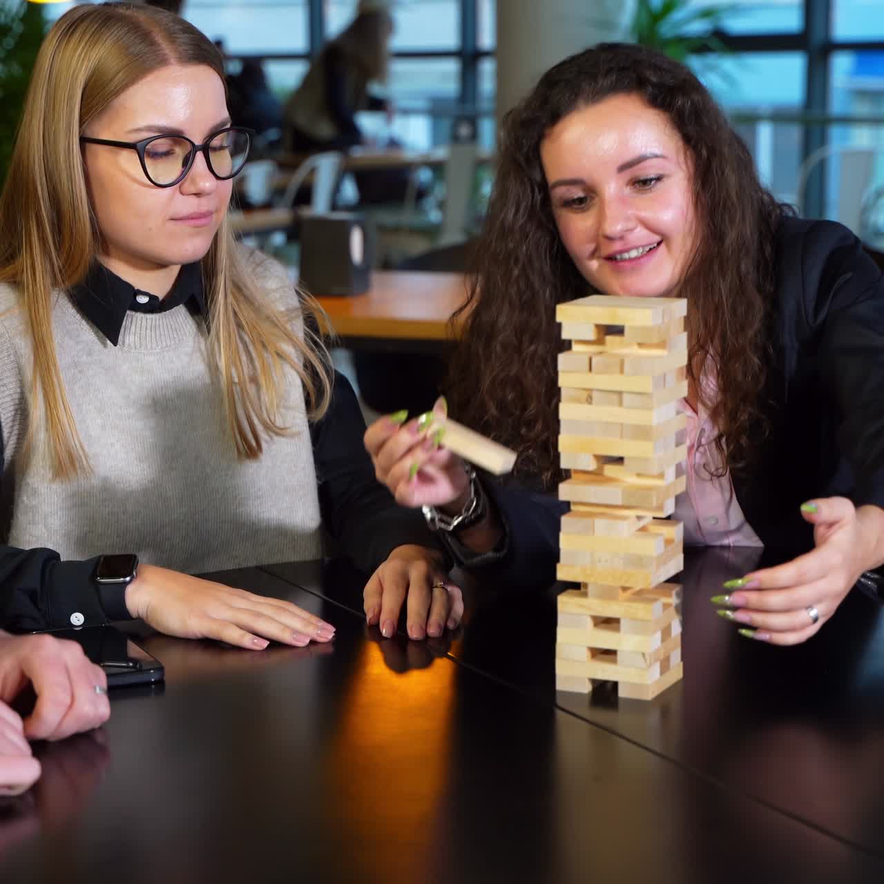 Cheerful happy group of young people play board game. Woman with long curly hair pulling brick from tower and other people watching her