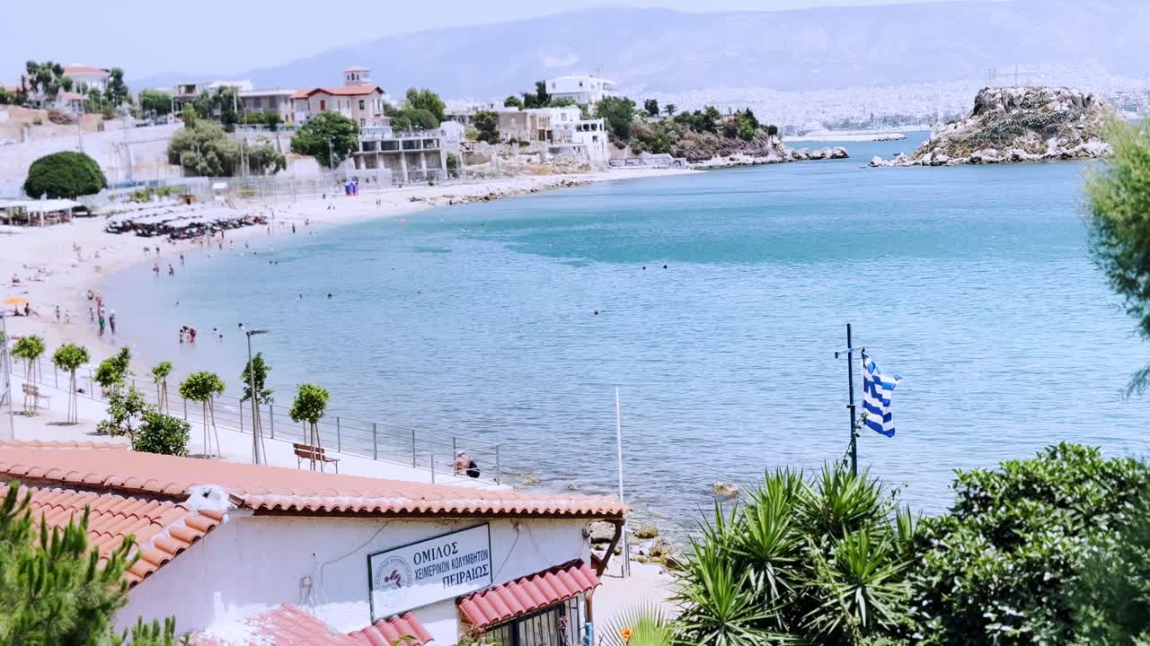 İnsanlar ve dalgalanan bayrakla güneşli bir Yunan plajından manzara.