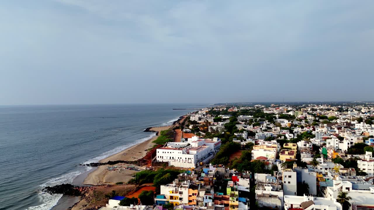 A high-angle drone shot reveals Puducherry's beautiful beach and sprawling urban area. The moody, overcast sky creates a dramatic scene of city meets sea