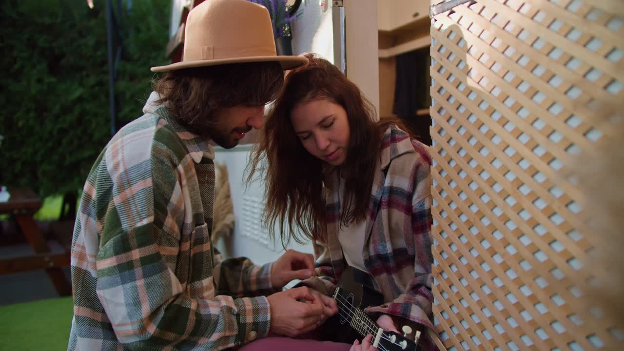 A brunette guy in a yellow hat helps his brunette girlfriend in a pink checkered shirt play a black ukulele while sitting near his trailer in a camp during a picnic outside the city in the summer