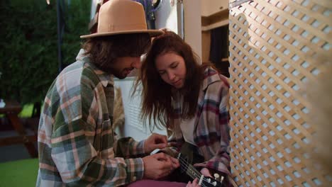 Ein-Brünetter-Typ-Mit-Gelbem-Hut-Hilft-Seiner-Brünetten-Freundin-Im-Rosa-Karierten-Hemd,-Eine-Schwarze-Ukulele-Zu-Spielen,-Während-Er-Im-Sommer-Bei-Einem-Picknick-Außerhalb-Der-Stadt-Neben-Seinem-Wohnwagen-In-Einem-Lager-Sitzt