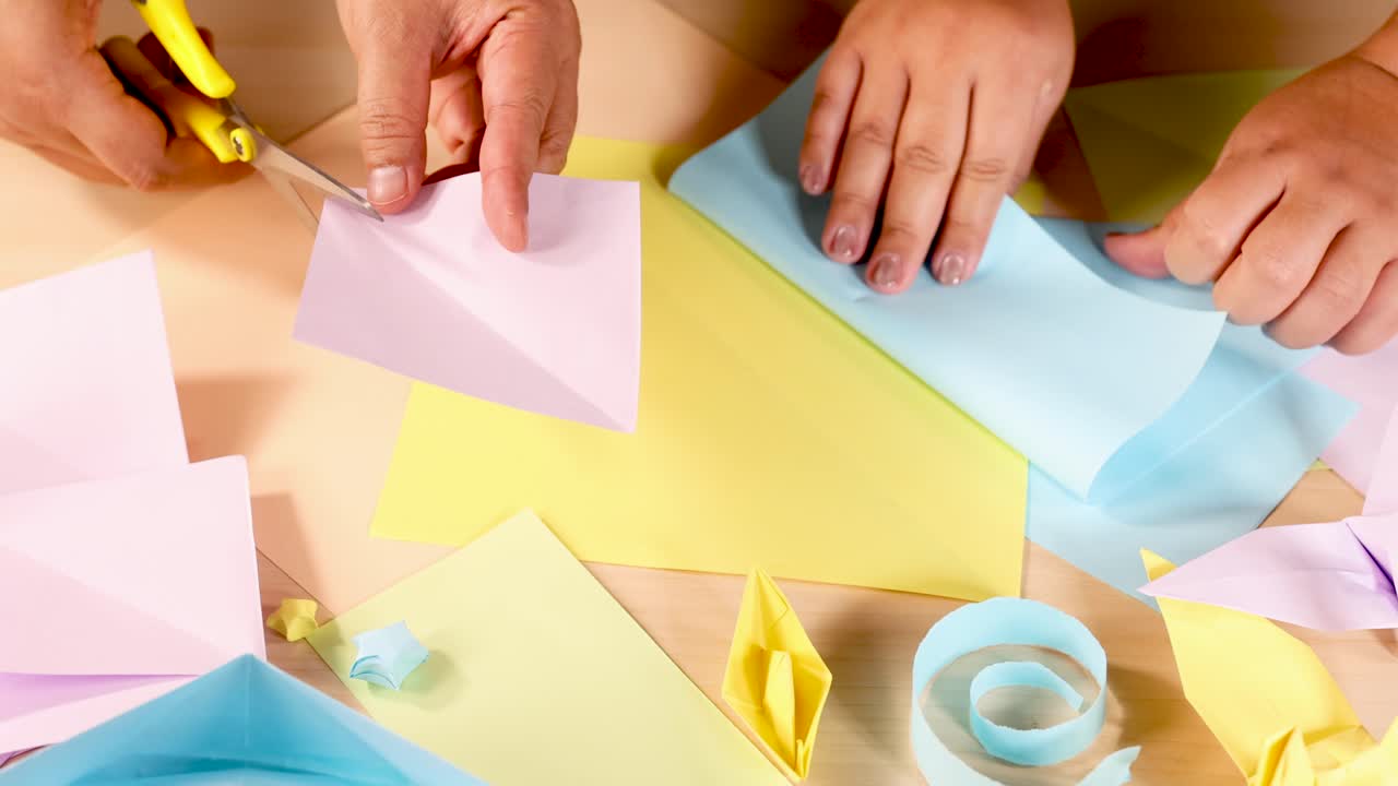 Two people cut and fold pastel origami paper on a table under bright, even lighting