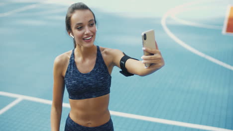 cheerful sporty woman with airpods smiling and showing biceps to smartphone camera while having videochat at sport court