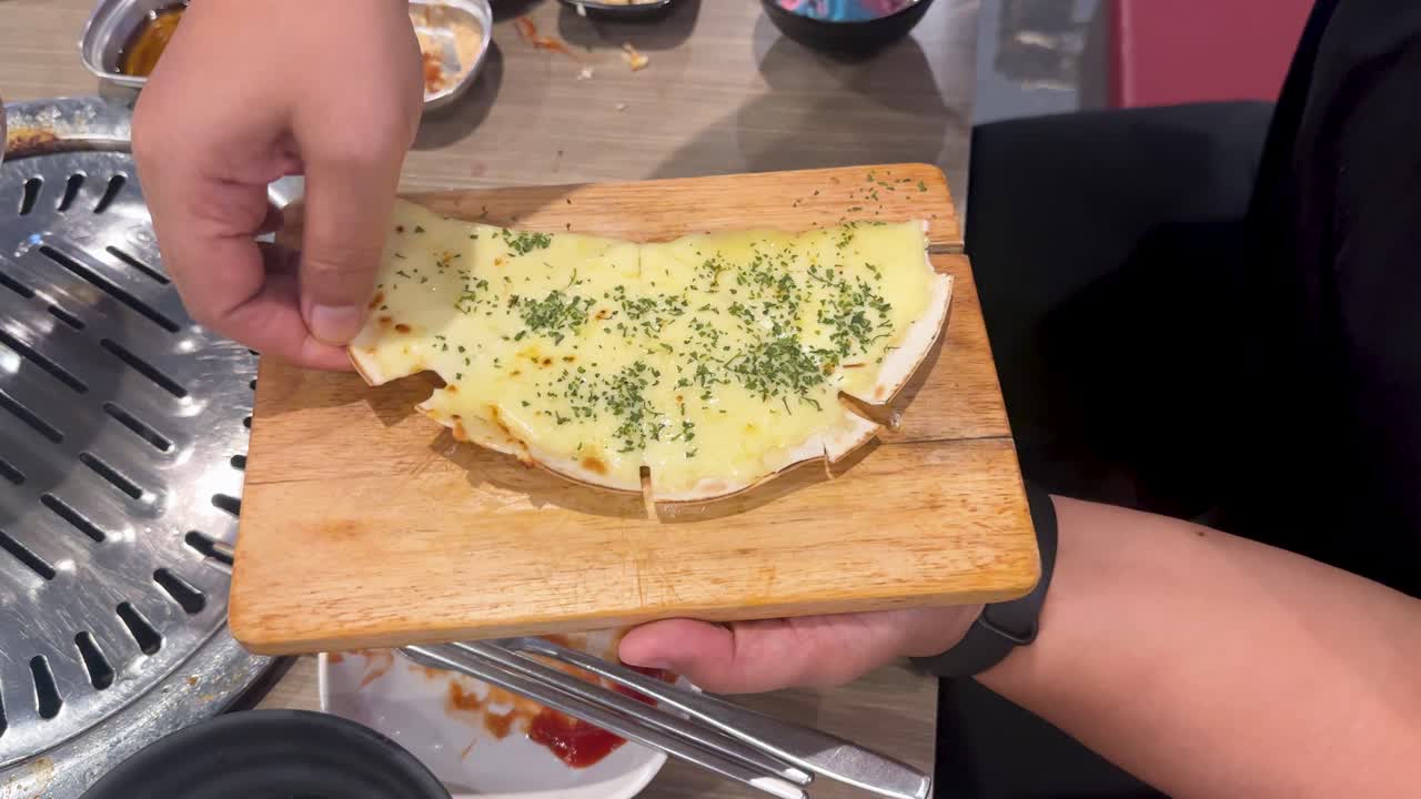 A hand stretches melted cheese over a dish in a Bangkok restaurant, highlighting a Korean BBQ setting with vibrant lighting