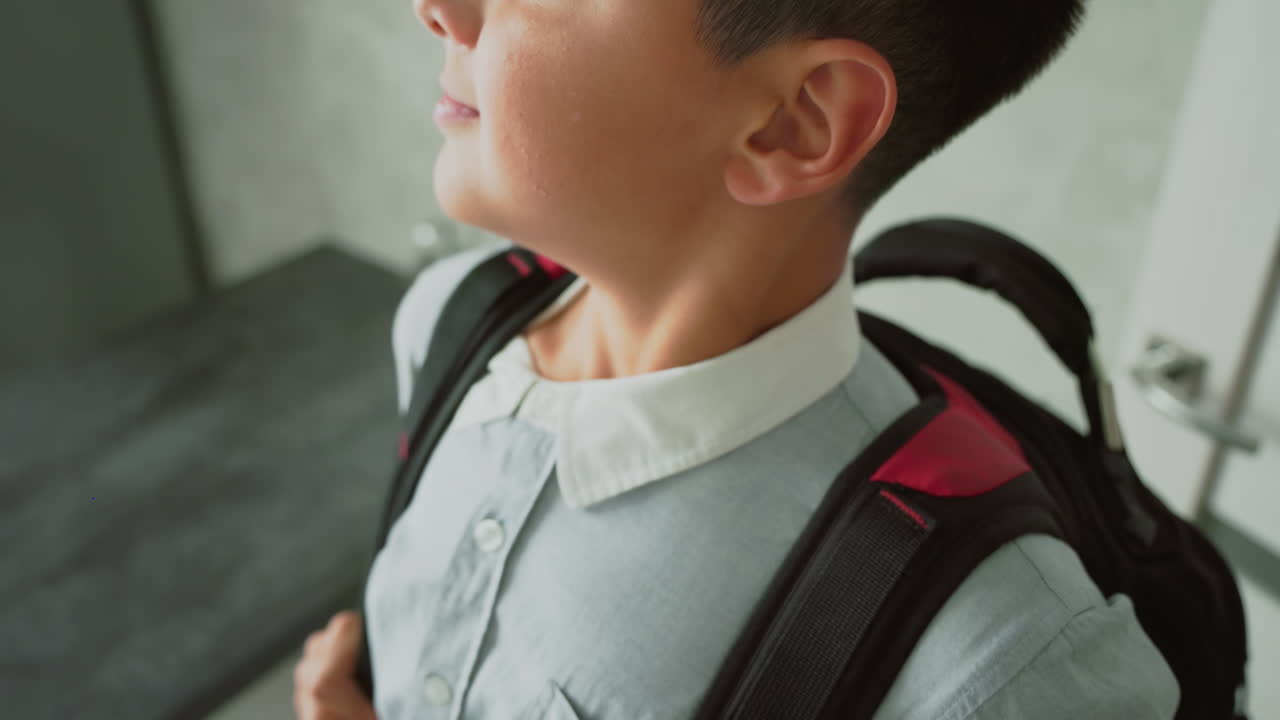 boy holding black school backpack by strap while wearing light button-up shirt, standing near kitchen counter, preparing for school day, showing focus, routine, independence, and start of daily journey