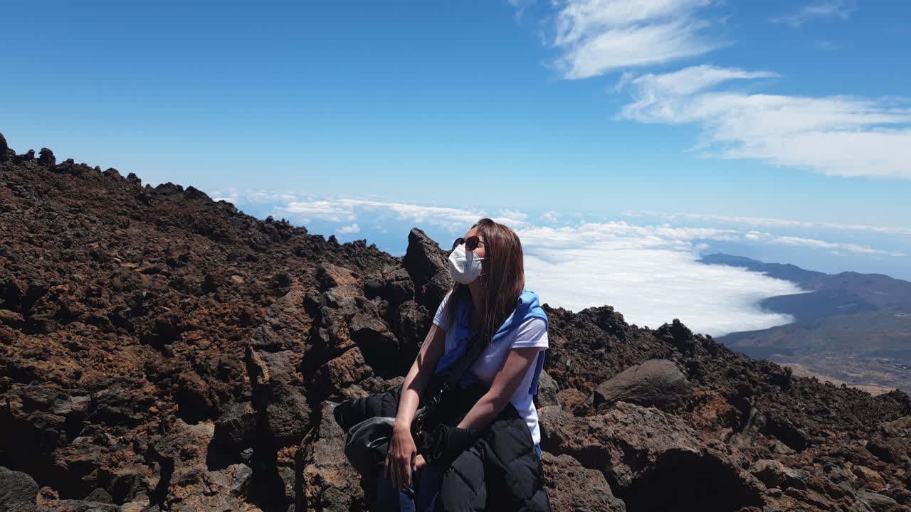 Slow motion video of a hiker wearing a mask resting on rocks at the summit of mount Teide in Tenerife national park, spain