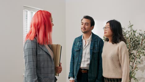 happy couple stand in empty room while inspecting apartment for rent with real estate agent woman and man look at place indicated by pink-haired girl touring rooms pointing out equipment appliances