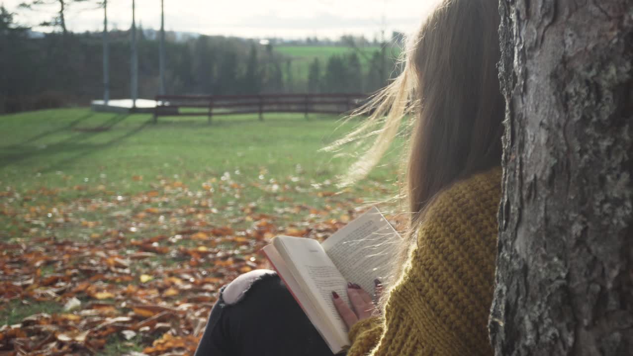 Adult girl is sitting under the tree reading a book in autumn scenery and relaxing