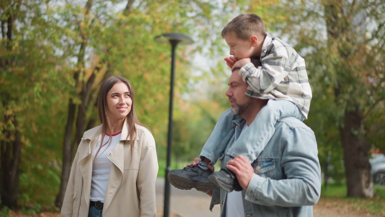 Primo piano di un uomo che porta un bambino sulle spalle mentre cammina con una donna in un parco all'aperto. Il bambino si tiene alla testa dell'adulto ed entrambi gli adulti sembrano essere coinvolti o osservare