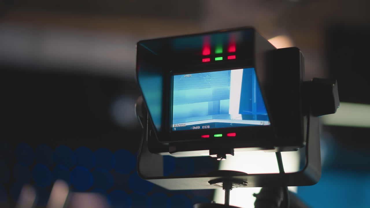 Close-up view of camera monitor showing broadcast studio setup, red and green indicator lights on, and professional TV production environment. Clear focus on blue-lit studio background