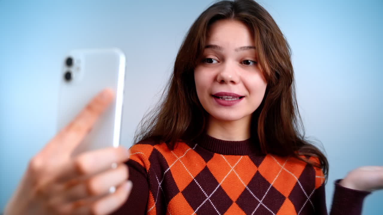 Young woman talking while filming herself on the phone and gesturing on a grey background