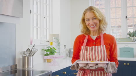 portrait of senior woman in kitchen at home holding tray of freshly baked cupcakes