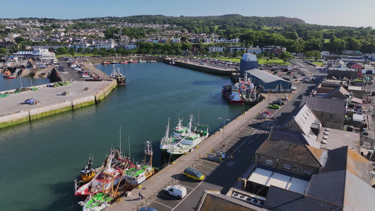 Howth Harbour, County Dublin, Ireland, May 2023. Drone slowly pushes along the quayside over moored Fishing trawlers with Howth Yachtclub and Seafront in the background on a bright sunny afternoon.