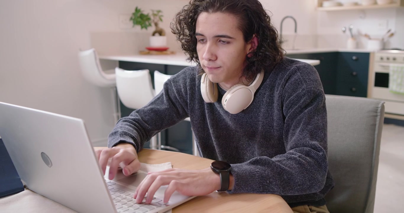 Homem na casa dos 20 anos cochilando no canto da cozinha, percebendo a tela do laptop, levantando a cabeça e retomando o trabalho digitando.