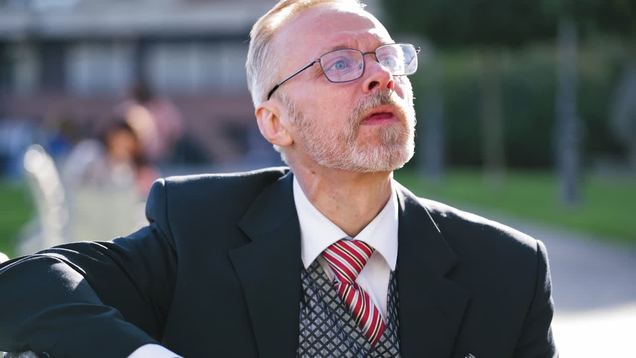Portrait of sad businessman in glasses outdoors. Mature man in tie and suit is upset about something while sitting with his phone in the street.