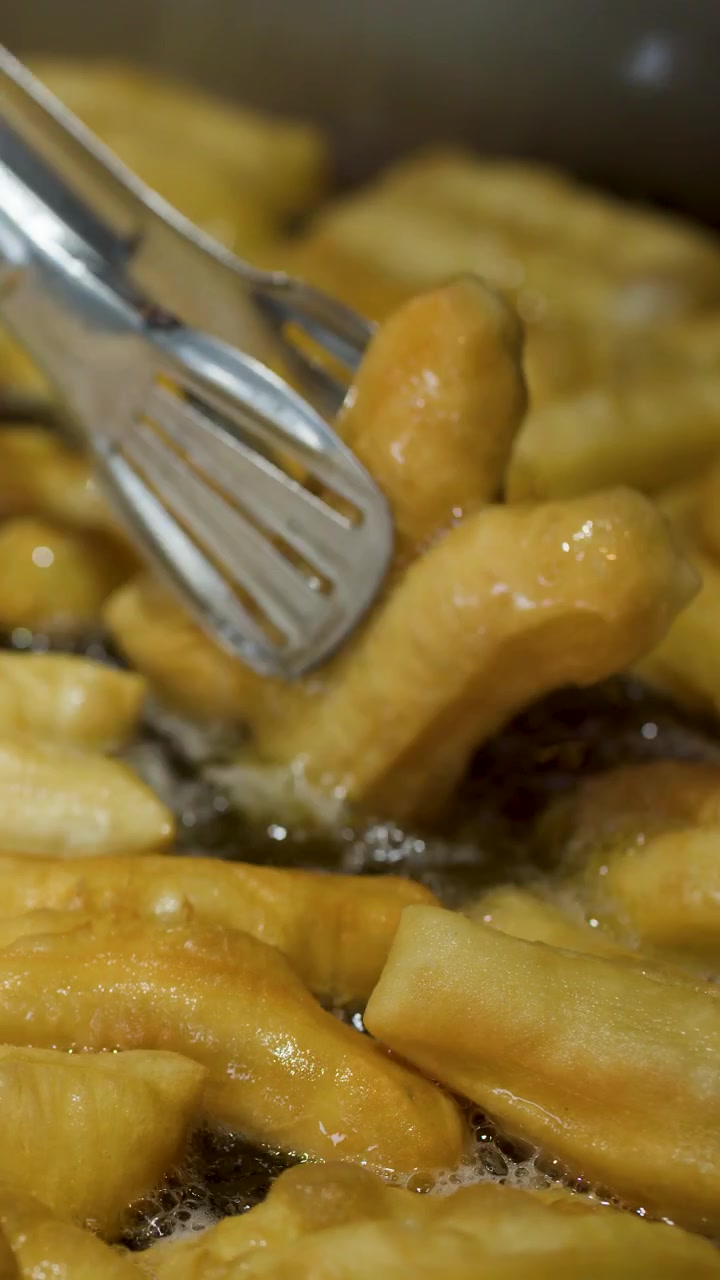 Primo piano di bastoncini di pasta dorati che friggono in olio bollente, girati con pinze di metallo.